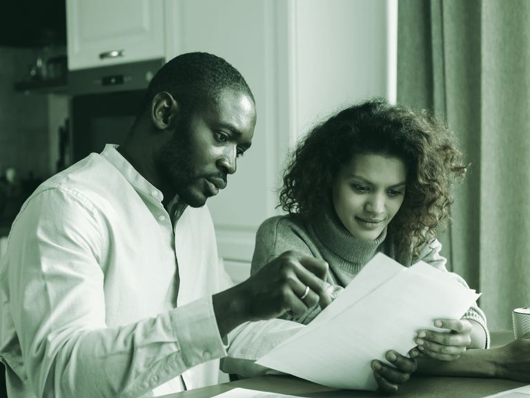 A couple at their kitchen table reviewing paperwork together on a laptop